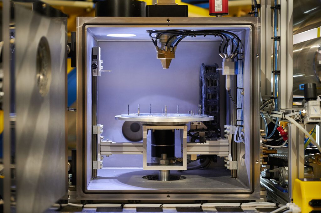 Blick in die geöffnete Versuchskammer. Auf der runden Trägerplatte in der Mitte sind mehrere in Schwerlosigkeit hergestellte kleine Metallzylinder zu sehen.
