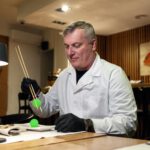 Ein Mann im weißen Kittel hält einen grünen kleinen Ball zwischen zwei Stäbchen.