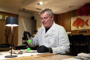 Ein Mann im weißen Kittel hält einen grünen kleinen Ball zwischen zwei Stäbchen.