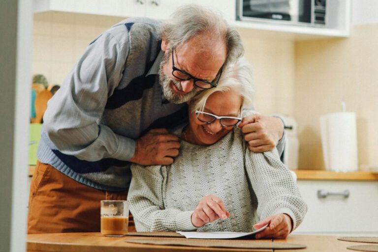 Ein älterer Mann hat die Hände auf die Schultern einer Frau gelegt, die vor ihm sitzt.