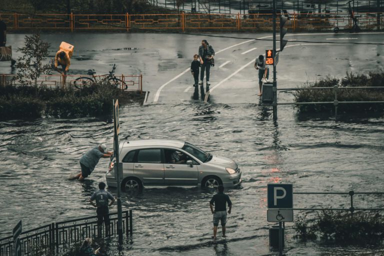 Eine überflutete Straße, ein Mann schiebt ein Auto durch das Wasser.
