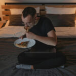 Mann sitzt abends auf dem Bett und isst Pizza