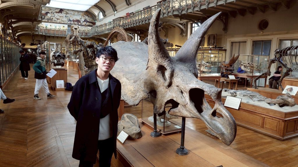 Ein junger Mann mit Brille steht vor dem Schädel eines Triceratops in einem Museum.