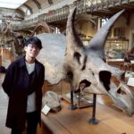 Ein junger Mann mit Brille steht vor dem Schädel eines Triceratops in einem Museum.