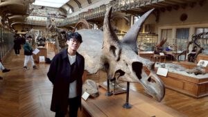 Ein junger Mann mit Brille steht vor dem Schädel eines Triceratops in einem Museum.