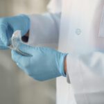 Eine Person im weißen Kittel hält mit zwei Händen, die in blauen Gummihandschuhen stecken, etwas.