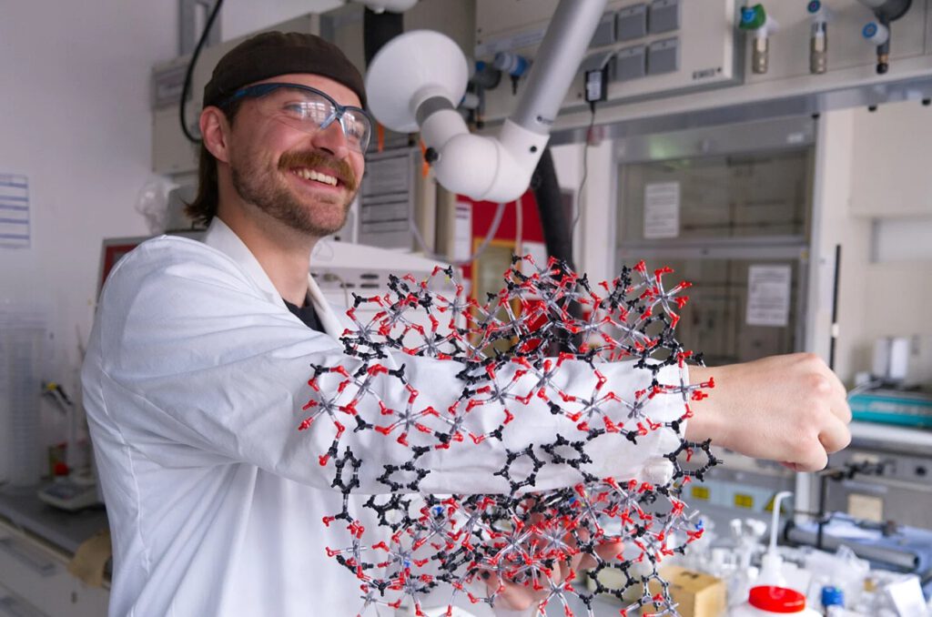 Kalle Mertin macht die Struktur von CAU-10-H anschaulich: Sein Arm passt durch das Modell der röhrenartigen Poren, in denen das Material Wassermoleküle aufnimmt und wieder freisetzt. © Christina Anders, Uni Kiel
