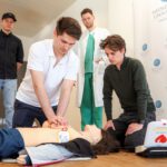 Mehrere Menschen stehen um eine Reanimationspuppe herum, während ein Mann eine Herzdruckmassage simuliert.