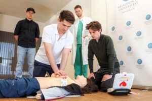 Mehrere Menschen stehen um eine Reanimationspuppe herum, während ein Mann eine Herzdruckmassage simuliert.