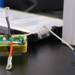 Die grüne Platine enthält den elektronischen Schaltkreis, mit dem die Forschenden Sensordaten über die vorhandenen Stromanschlüsse einer Batteriezelle übertragen. Die Batteriezelle befindet sich im Hintergrund. © Christina Anders, Uni Kiel