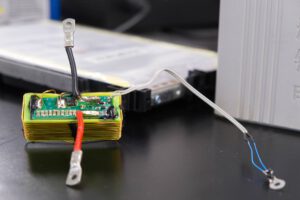 Die grüne Platine enthält den elektronischen Schaltkreis, mit dem die Forschenden Sensordaten über die vorhandenen Stromanschlüsse einer Batteriezelle übertragen. Die Batteriezelle befindet sich im Hintergrund. © Christina Anders, Uni Kiel